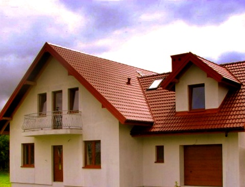 Dwupiętrowy dom z czerwonym dachem z dachówki, białymi ścianami i balkonem z metalową balustradą, garaż z brązową bramą.